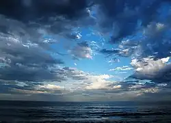 Late daytime view looking far out over an ocean from a beach, which is out of view off the bottom margin. Three-fourths of the shot features a sky marked by heavy cloud cover, which is parting near the middle, revealing a dazzlingly bright cerulean blue sky that darkens near the margins. The ocean is striated with waves coming in parallel to the horizon.