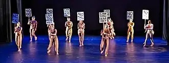 Dancers in brown facing the audience, holding handwritten black-and-white signs saying "no justice," "no peace," and "can you hear me?"