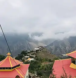 Picture of houses in a valley between two treed mountains. Clouds in the sky. Buildings with red roofs in the foreground.