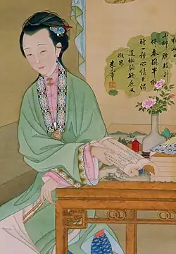 A painting of a woman seated at a desk with a red snuff bottle, a scroll, and a vase with flowers