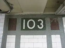 Tiles on the walls of the platform extension. There are vertical and horizontal green mosaic bands, surrounding white ceramic bricks on the bottom and three rectangular mosaic boxes on the top. The center box contains the number "103" in white tiles on a green background. The left and right boxes contain bronze tiles.
