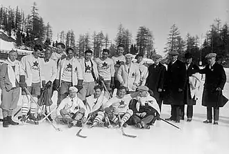 Black and white photo of a hockey team outside on natural ice, including fourteen players dressed in hockey equipment white sweaters with a maple leaf crest, and four men dressed in dark suits and overcoats