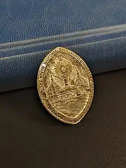 A commemorative pin from the 1941 Eucharistic Congress. Depicted are the Cathedral of Saint Paul and the Basilica of Saint Mary, in between them the Tissot monstrance. Surrounding is the text "THE NINTH NATIONAL EUCHARISTIC CONGRESS. SAINT PAUL JUNE 1941 MINNEAPOLIS"