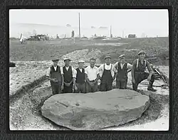 Seven men wearing work gear, including flat caps, pose behind a stone that they have excavated. The stone must be over 10 foot long, and has been exposed laying down.