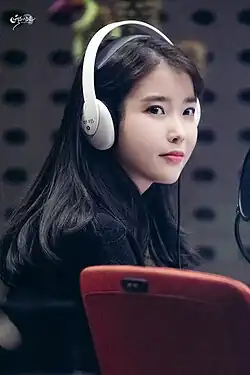 Woman with long black hair wearing white over-ear headphones and a black outfit sitting in a recording studio with her eyes looking slightly to the side, with a red chair visible in the foreground.