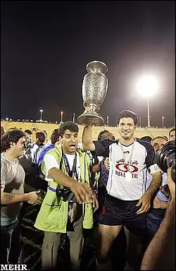 Photograph of one player holding the trophy above his head and smiling at the camera while standing among a crowd of other players and officials