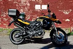 Black and yellow BMW F800GS with topbox and motorcycle helmet on the seat, parked in front of a brick wall covered in peeling red paint