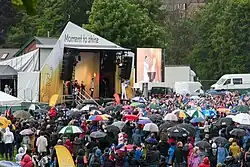 A crowd under umbrellas watches a man on a stage light a cauldron from a torch