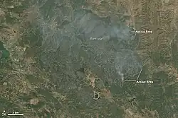 The Rocky Fire is seen from directly above as a very roughly circular brown patch on green-to-tan land cover, with the top and left sides issuing gray smoke