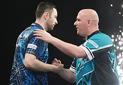 Luke Humphries (left) shakes hands with Rob Cross at a Premier League Darts event, with a pyrotechnic in the background. Rob places a hand on Luke's shoulder.