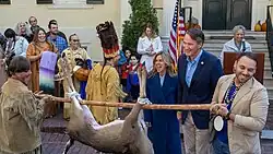 An older white man in a dark blue blazer smiles as he is presented with a dead deer hanging upside down held by two men in contemporary Native American attire.