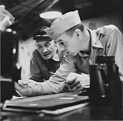 Black and white photograph of two men in naval uniforms examining some documents placed on a table