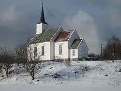 The picture depicts the Skålvær Church. It is a small building with its walls painted white and a darker roof. The area around the church is covered in snow and the trees have no leaves.