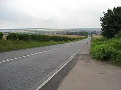 From camera position at right side of a straight road looking down a steep hill with hedged agricultural fields on both sides and town in hazy distance at river level