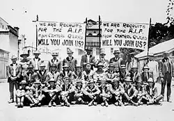 Black and white group portrait of a group of men wearing suits seated and standing in front of two banners. Both banners read "We are the recruits for the A.I.F. from Central Q'Land [Queensland] will you join us?" in block letters.