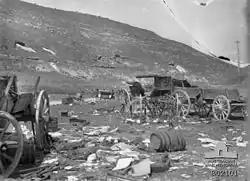 Cart wheels, wagons destroyed barrels and other wreckage in foreground, two soldiers on the road in the middle distance near a staff car