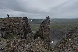 A hiker stands on a mountain summit near a rocky outcropping, with a vast landscape with lakes in the background