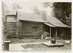 A large log cabin-like building, with a chimney, window with curtains, a small porch, and a water pump out front. The building is rectangular with a round room in the back