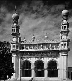 A black and white image of a mosque with three arched entrances, flanked by two minarets on the sides.