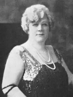 A white woman with light-colored hair in a curly set style, wearing a sleeveless gown and pearls