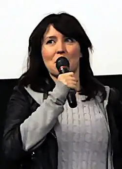 blurry photo of Alice Lowe wearing a grey sweater and black jacket, speaking into a microphone, looking right of camera