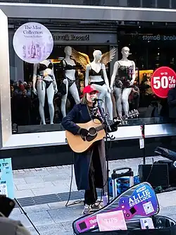 A teenage girl plays guitar by a microphone on a street