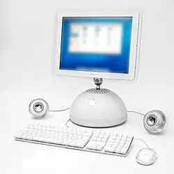 A white computer with a hemispherical base and shiny Apple logo, attached to a flat screen with a metal arm. A keyboard and mouse sit in front of the computer, while flanking the base are round, shiny speakers.