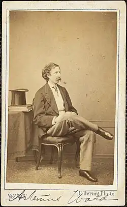 Photograph of Artemus Ward, sitting with his right leg crossed over his left, a top hat and books sitting on the table to his right. A typed caption at the bottom of the image reads "H. Hering Photo" and his name is written at the bottom.