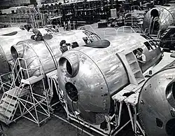 A black and white photograph of an aircraft factory. 1 female and 2 male workers are seen working on 3 front bodies of aircraft.