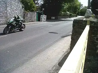 Racing motorcycle rider on left adjacent to a high wall approaching camera position with road stretching out behind to bend in distance