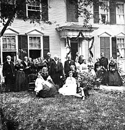 A group of people standing and sitting outside a New England-style tavern in 1868