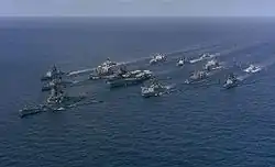 A large collection of ships sailing on the sea from the back right to the front left. At the center of the cluster of ships is an aircraft carrier, with a battleship in front of the carrier. Other ships of various types are sailing in a roughly circular formation to provide defense for the aircraft carrier.