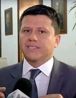 A man in his forties stands in front of a microphone wearing a business suit. He had tan skin and short black hair. There are portrait paintings visible in the background.