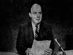 black and white photo of a man sitting at a desk holding a sheet of paper and speaking into a microphone