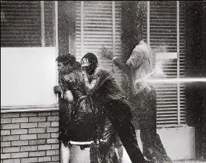 Three black high school students, two boys and a girl, facing into a storefront window to avoid being hurt by a water cannon blasting a boy at his back; all three are dripping with water