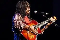 alt=A man with hair braids is wearing sunglasses and playing a guitar. Milton Nascimento