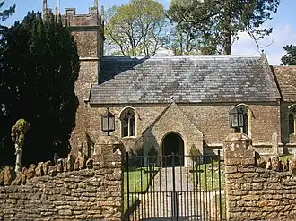 Small stone building with square tower partially obscured by trees. In the foreground is a stone wall with metal gates.