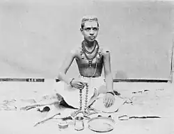 This image shows a young Brahmin Priest sitting on floor with folded feet, "tilak" on his forehead, rudraksh mala around his neck, mala in his hand and other Pooja ritual necessities laying around him.