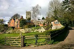 Red brick buildings in front of grey stone church. In the foreground in a grassy field contained within a hedge and fence.