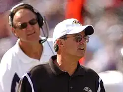 Candid photograph of Zauner standing in front of Brian Billick on a football sideline wearing a black polo shirt and white baseball cap both of which bear the Baltimore Ravens logo