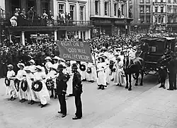 A procession of Suffragettes, dressed in white and bearing wreaths and a banner reading "Fight on and God will give the victory" during the funeral procession of Emily Davison in Morpeth, Northumberland, 13 June 1913. Crowds line the street to watch.