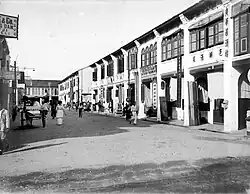 High angle view of a street lined with two and three-story structures. The street is filled with crowds, including horse-drawn and hand-pulled carriages.