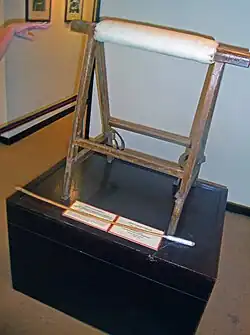 A wooden stand of two triangular portions with a padded brace between them on a black pedestal. A hand points from the left, and a small wooden stick with cotton wrapped around one end is in front.