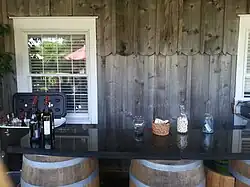 A black marble bar with wine bottles sitting atop oak barrels, with a wooden wall and white-colored window in the background.
