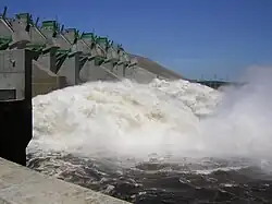 Image of a wall like structure in left, a raging river in the right and power lines located on low hills