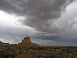 Dark, rolling storm clouds lower over a desert landscape; a butte stands in the near distance, left of center.