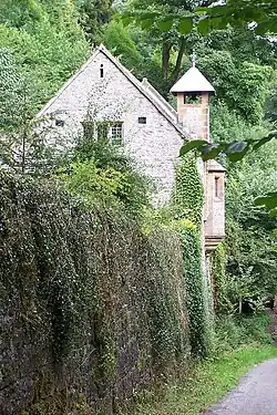 Beyond a tall wall is the end of a chapel with a triple window in the gable, a shorted tower to the right with a pyramidal roof, and an oriel window protruding more to the right