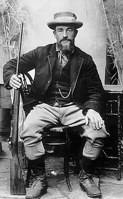 Rugged, bearded, hatted man in Victorian clothing seated holding a rifle in the right hand.