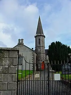 Behind a wrought iron gate sits a church with grey stone quoins. An octagonal steeple with a tall pointed roof adjoins the right side of the nave and contains a ground-level entry with a red door.