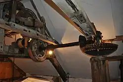 A detailed view of another mechanism, the sack hoist. This is in the grain loft above the first floor, and is designed to be driven by the vertical shaft as needed. On the right of the picture is the top of the vertical shaft that has passed through the height of the whole building, capped with a small iron bevel gear. This is mated with another bevel gear on a short horizontal iron shaft that transfers the drive to a wooden wheel suspended below a joist. A canvas strap around this wheel can be tightened or slackened and forms a clutch, to transfer the drive to another wooden wheel above the joist as required. The higher wooden wheel drives a wooden drum around which a chain is wound. Sacks can be lifted through the floors on this chain when the canvas clutch is engaged, or lowered controllably by allowing it to slip a little. The space is low, and angled under the pitch of the roof. A modern white-painted lining has been fitted under the roof, but the joists and rafters in bare pale wood can still be seen.